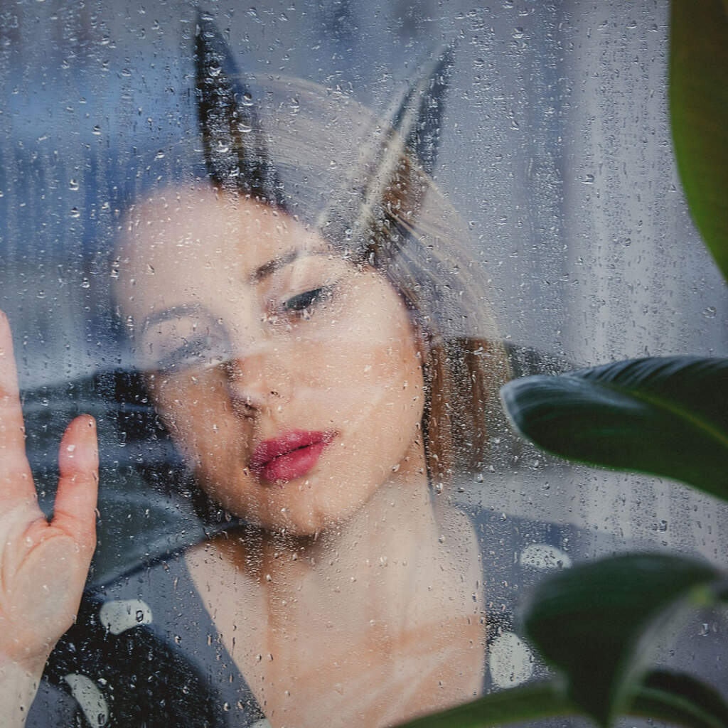 young sad woman near wet window after the rain misses the ficus